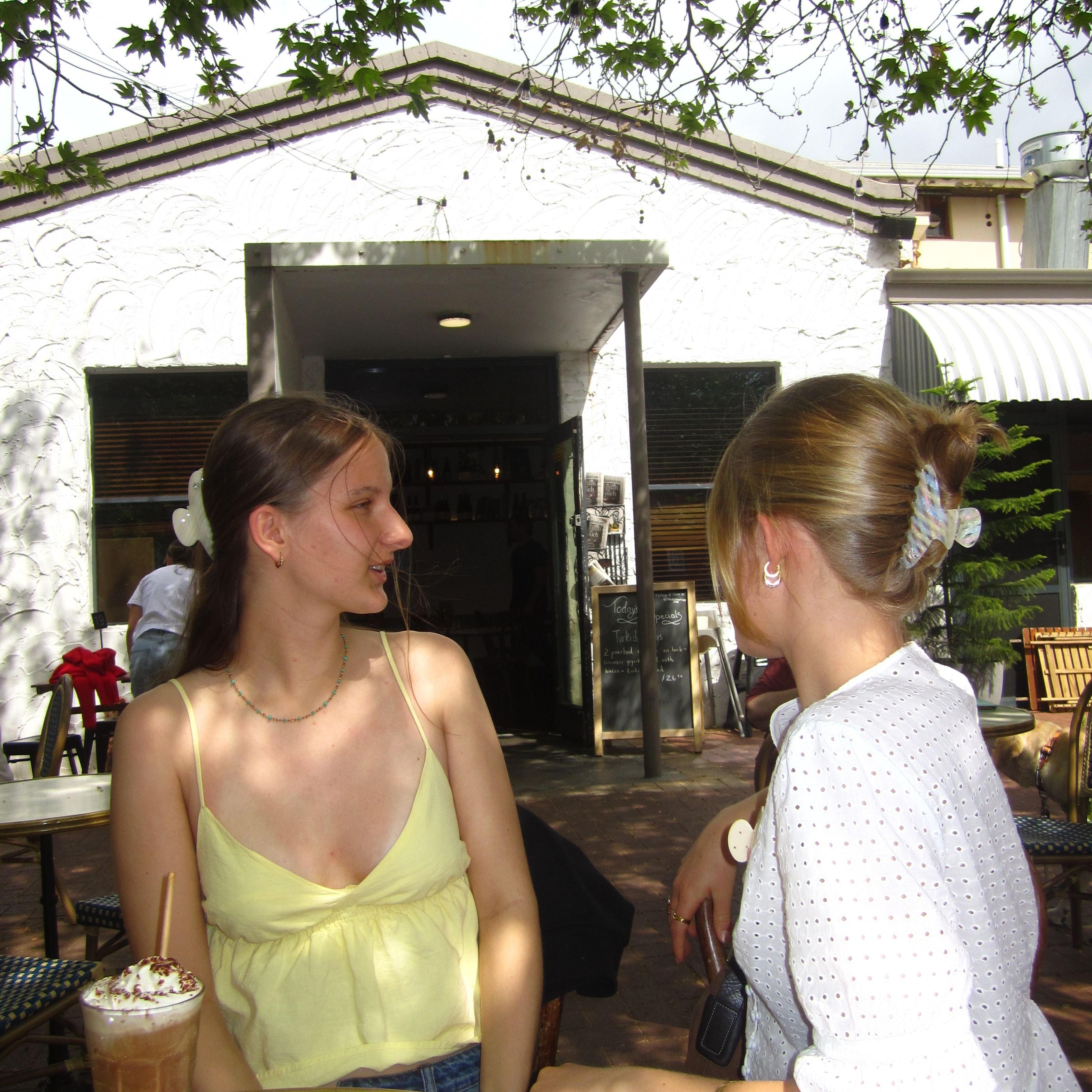 Two Luna Wave women sitting outdoors, one holding a drink, with a building and trees in the background wearing initial claw clip K