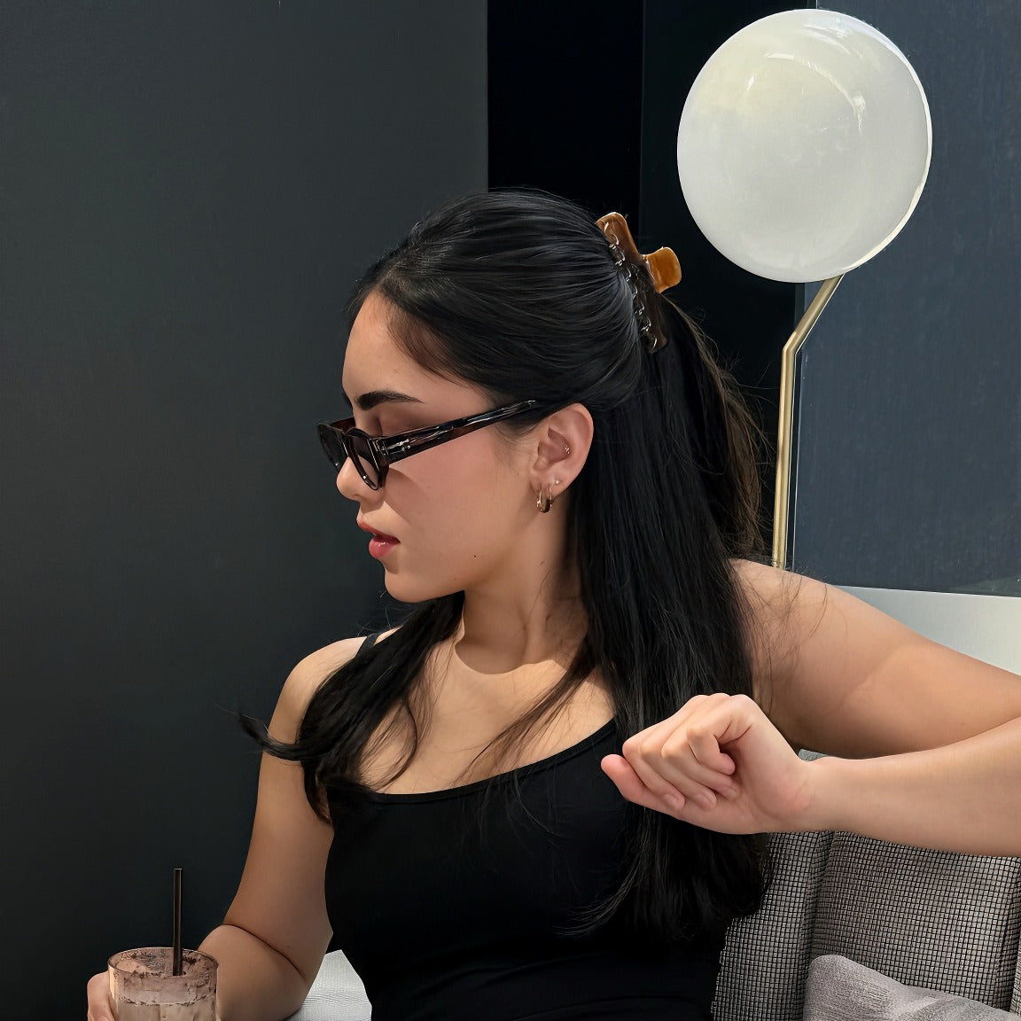 Model wearing Luna Wave Jelly Claw in Amber in a half up hairstyle while seated on a sofa holding a drink.