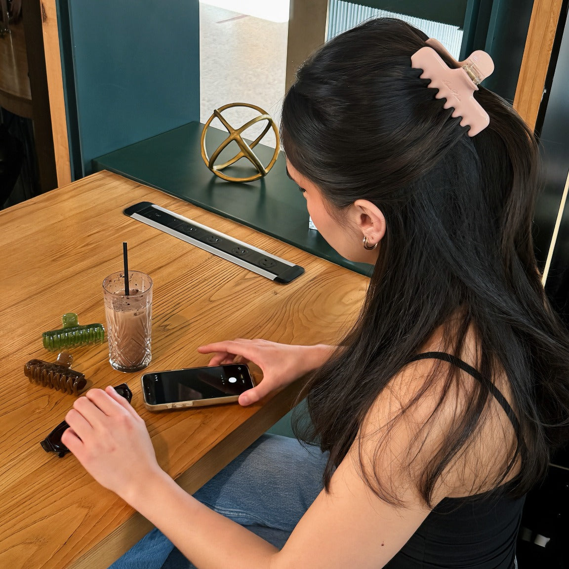 Rear side view of a model wearing the pink Luna Wave Jelly Claw in Rose in a half up hairstyle at a cafe table.