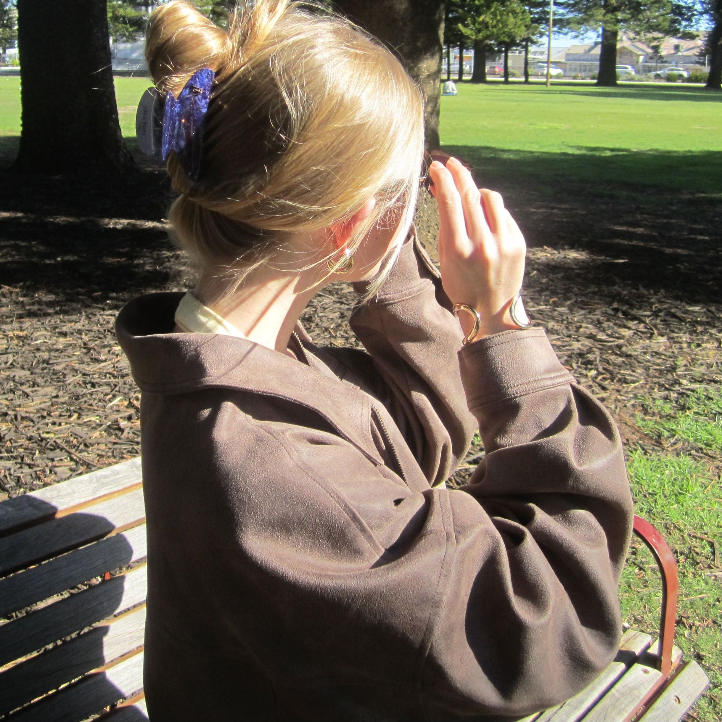 A lady sitting on a bench in a park, holding a sun glasses on her face and wearing a Luna Wave midnight galaxy claw
