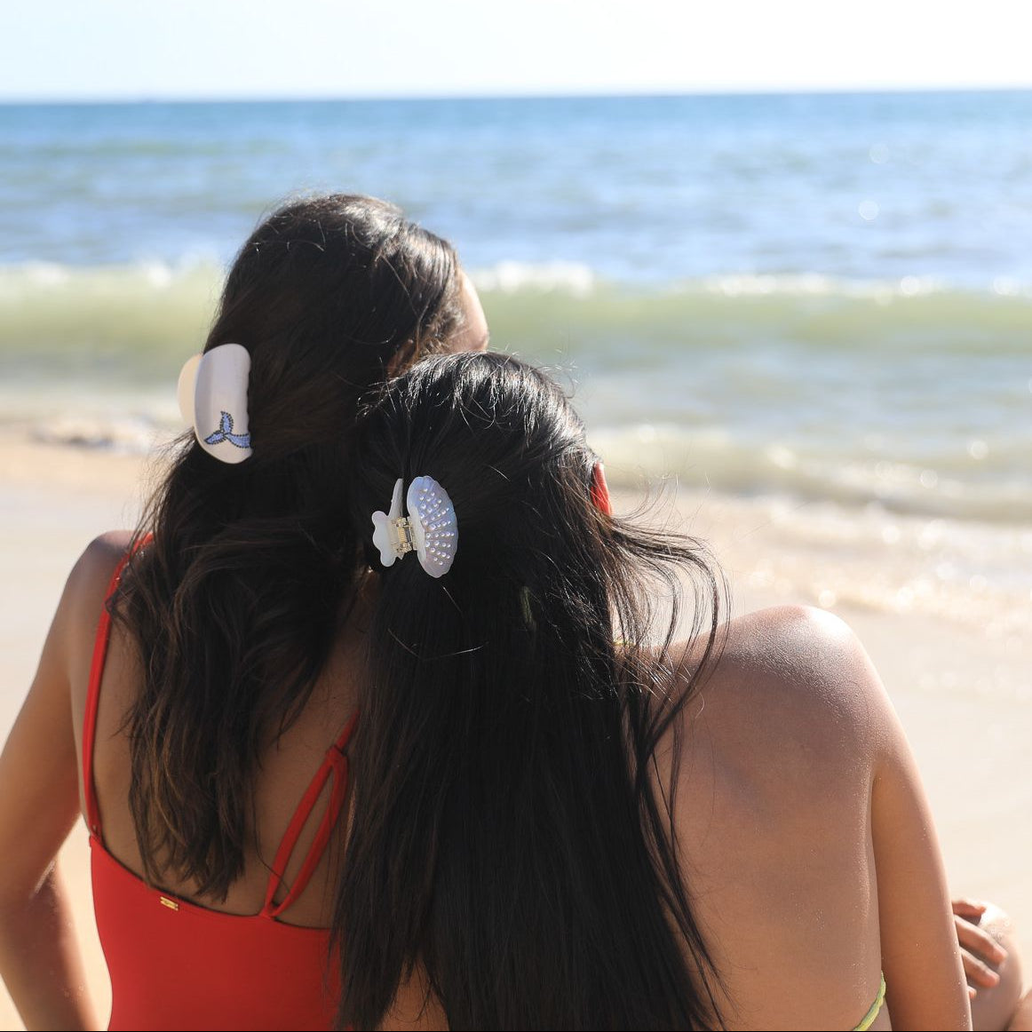 Two models sitting on a beach with ocean view, wearing luna wave whale bubbles and white aurelia hair claw clips.