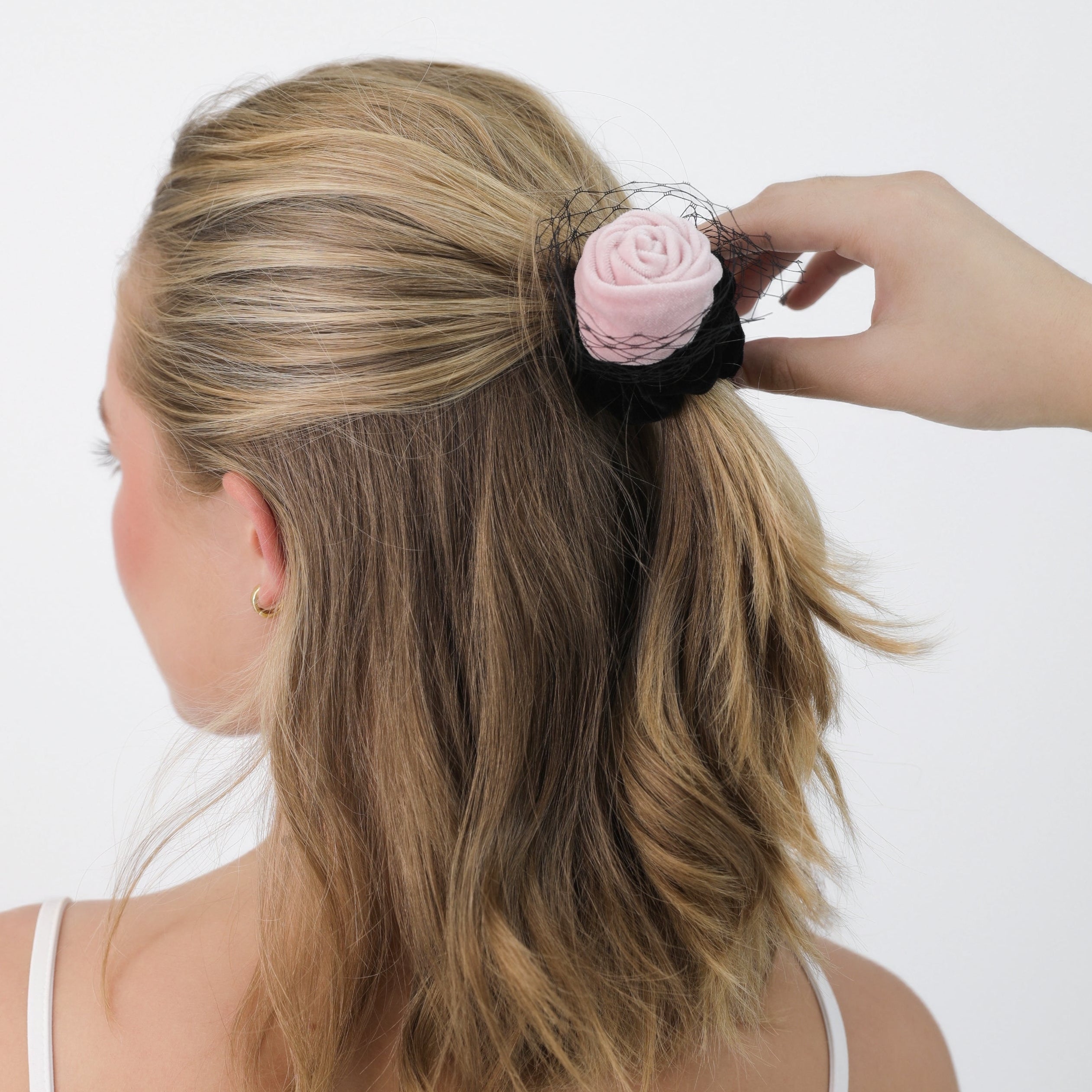 Woman adjusting a Luna Wave pink amelie rose scrunchie in her blonde hair against a white background